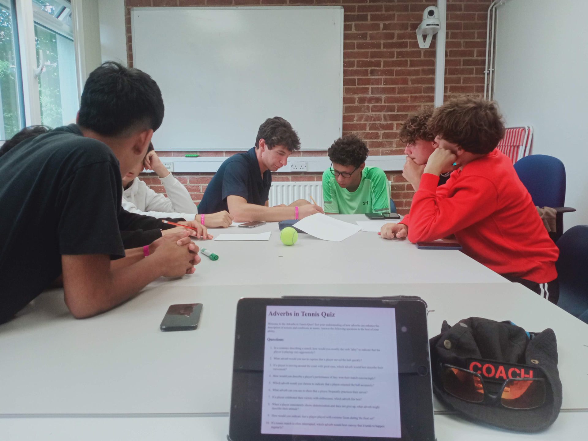 Students working on a tennis English quiz around a classroom table with a coach